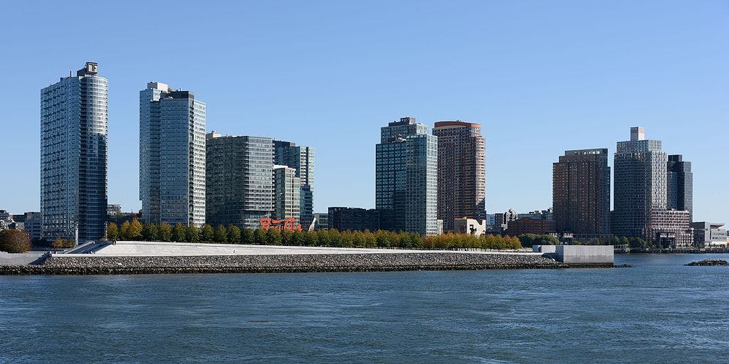 A row of modern high-rise buildings stand behind a waterfront park lined with trees on a clear day.