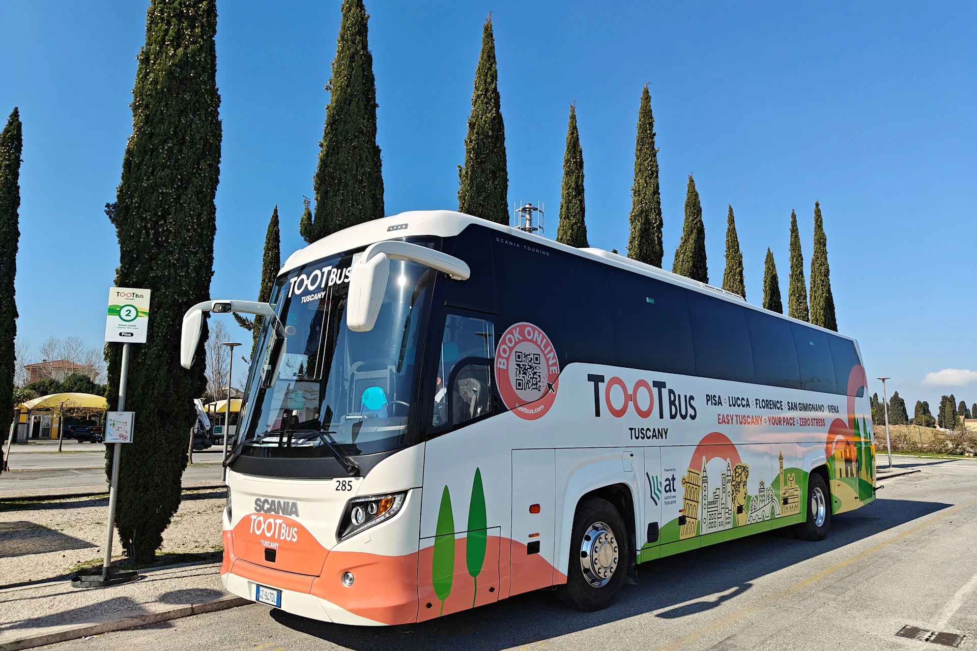 A Tootbus Tuscany sightseeing bus parked on a sunny day, with tall trees and clear blue sky in the background.