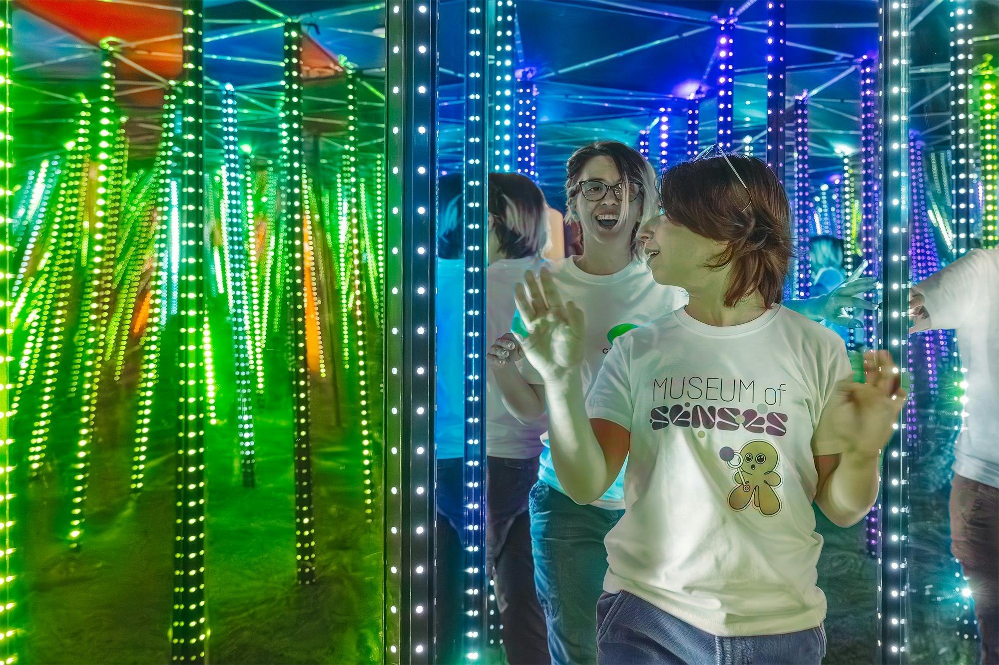 Two people in a mirror maze with colourful LED lights wearing "Museum of Senses" T-shirts, smiling and exploring.