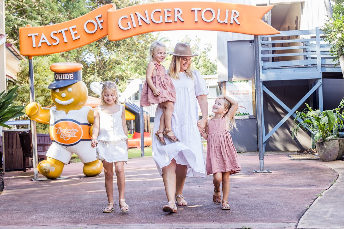 A woman in a white dress and hat walks with three young girls under an orange "Taste of Ginger Tour" banner near a gingerbread man statue.