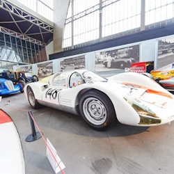 En vit racerbil med "148" på sidan visas tillsammans med flera veteranbilar i ett väl upplyst inomhusmuseum.