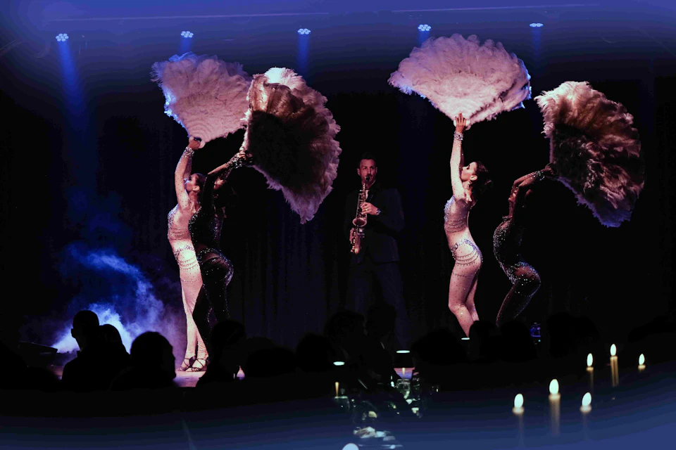A saxophonist performs on stage surrounded by dancers holding large feather fans, with an audience visible in silhouette.