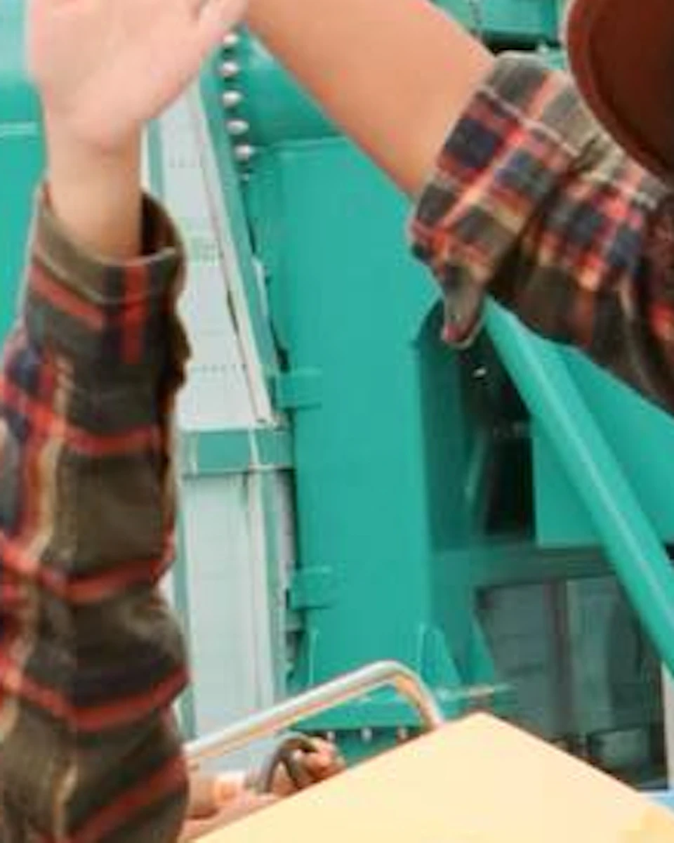 Two people in plaid shirts raise their hands and smile on a carnival ride, surrounded by colorful decorations and palm trees.