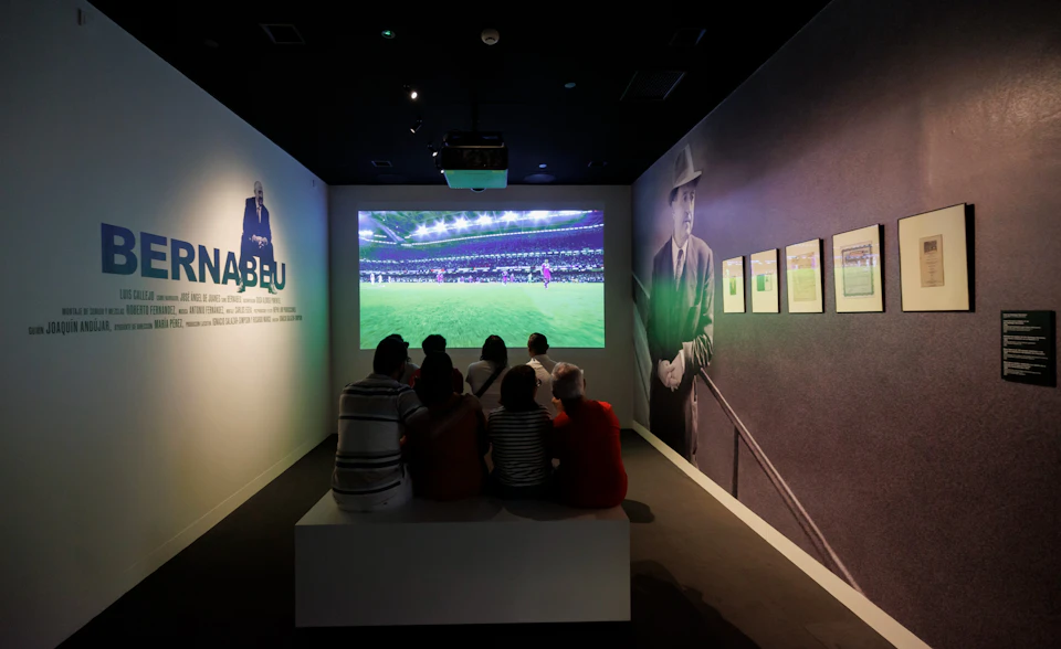A group of people watching a soccer match on a screen in a dim room with images and text on the side walls.