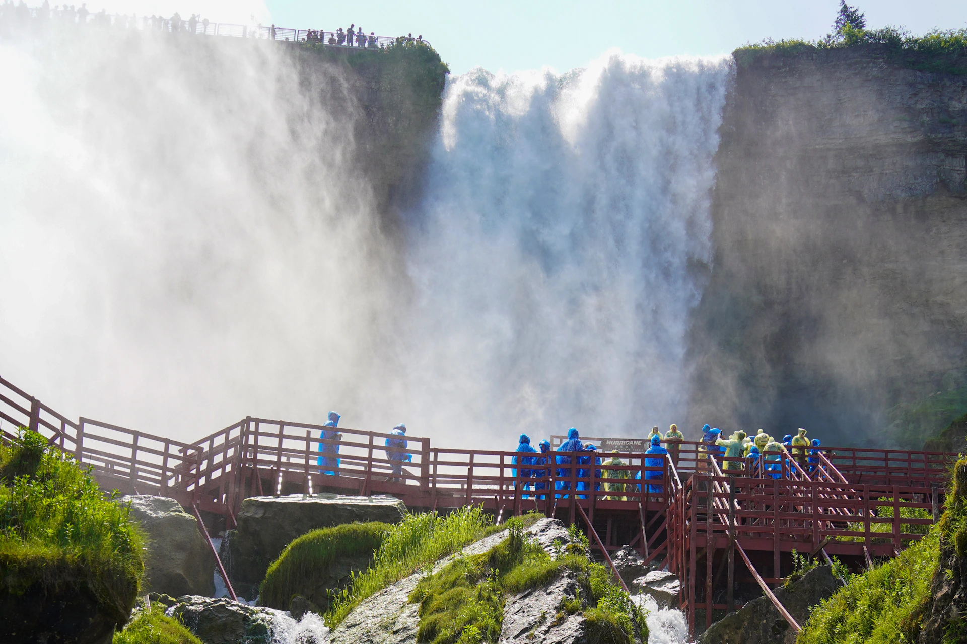 People in blue rain ponchos on a wooden walkway near a large waterfall, with mist and greenery surrounding the area.