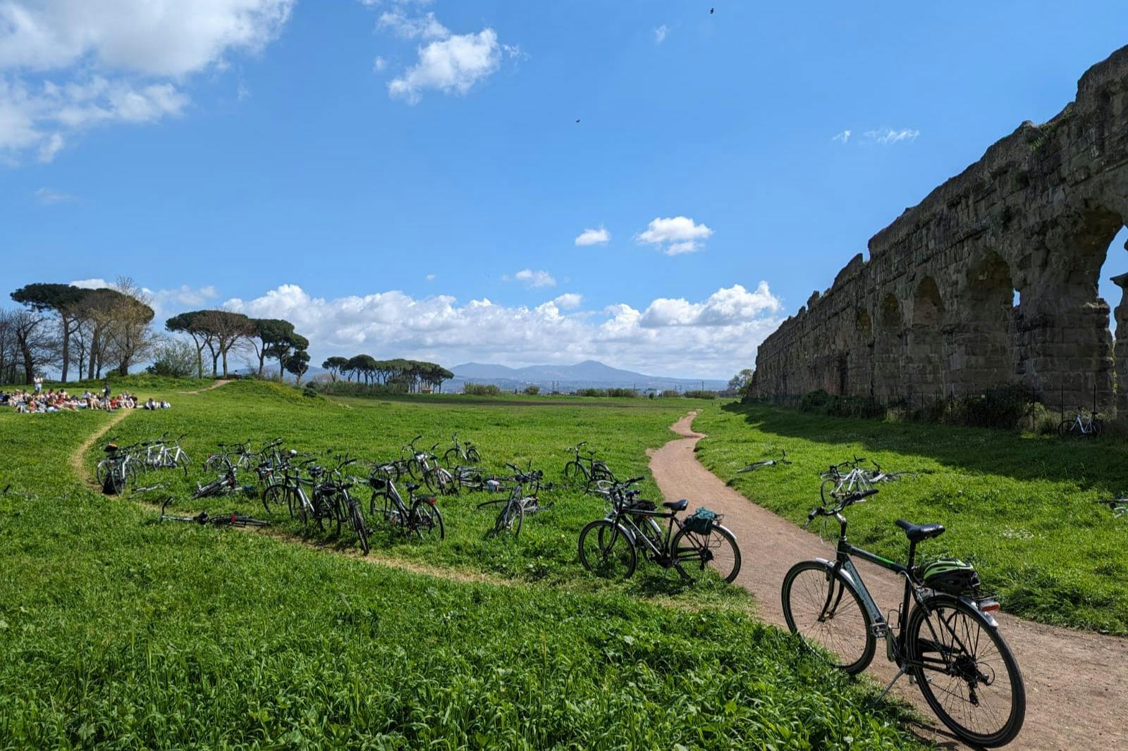 A grassy field with several bicycles parked on the ground, a dirt path, and ancient ruins under a blue sky with clouds.