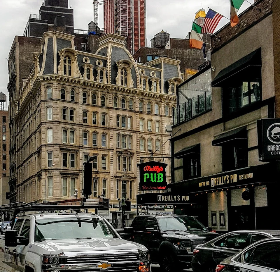 A street in a city with parked vehicles, a historic building, and flags flying near O'Reilly's Pub.