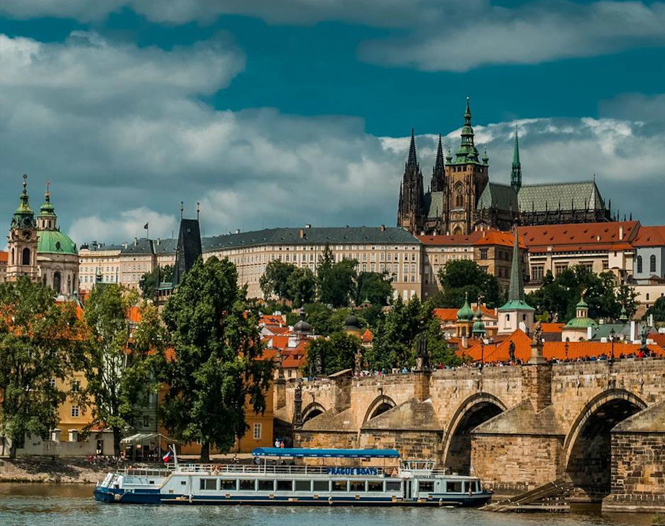 Historic European cityscape with a river, stone bridge, tour boat, buildings with red roofs, and a cathedral with spires.