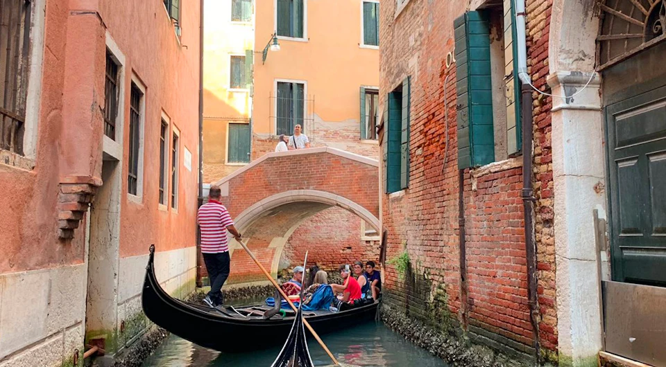 A gondolier navigates a canal with a group of tourists in Venice, passing under a stone bridge, surrounded by colorful buildings.