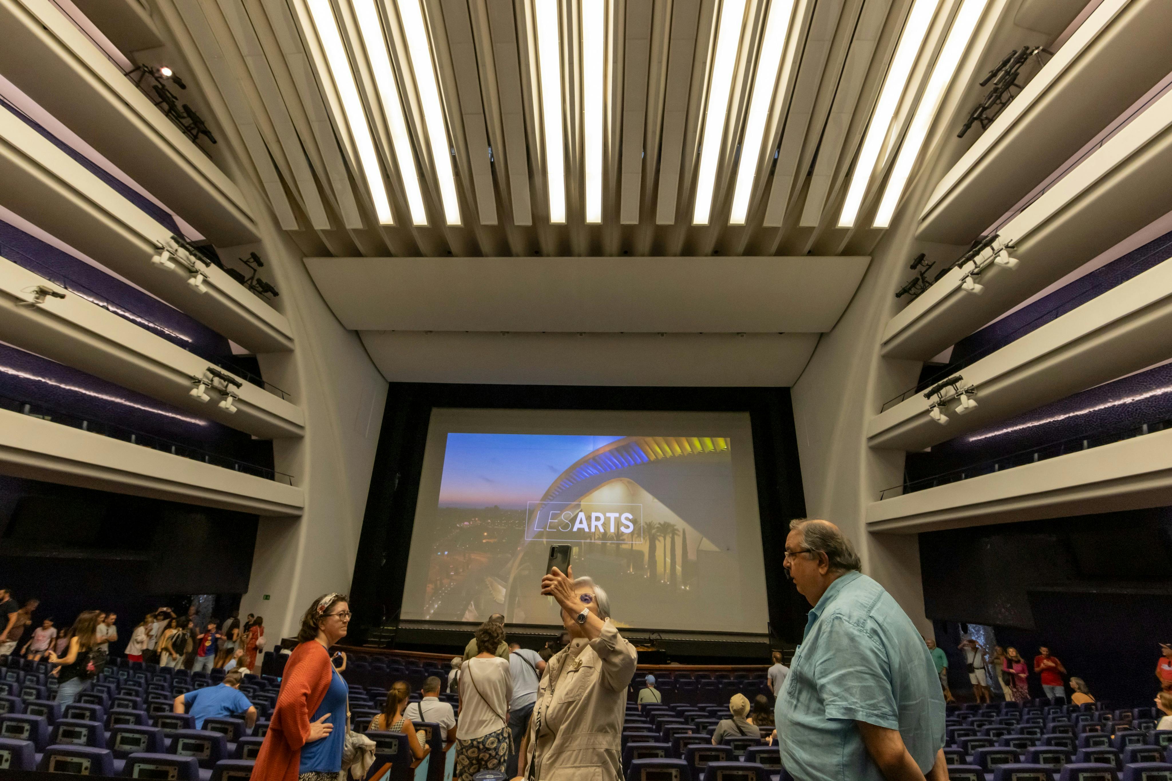 Menschen in einem Theater mit Reihenbestuhlung, die vor einer großen Leinwand mit der Aufschrift "LES ARTS" Fotos machen.