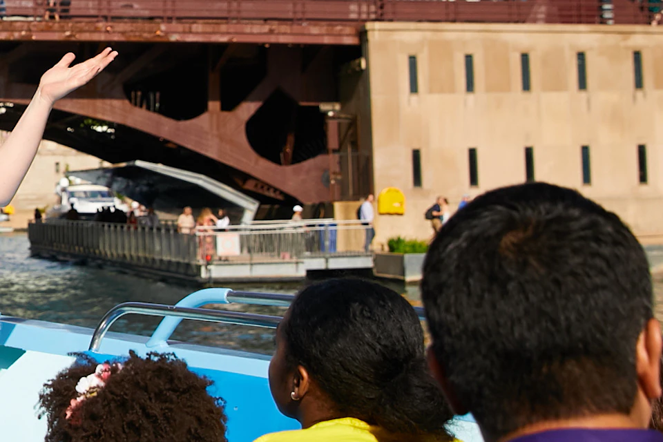 A tour guide speaks to passengers on a boat with city skyscrapers and a bridge in the background.