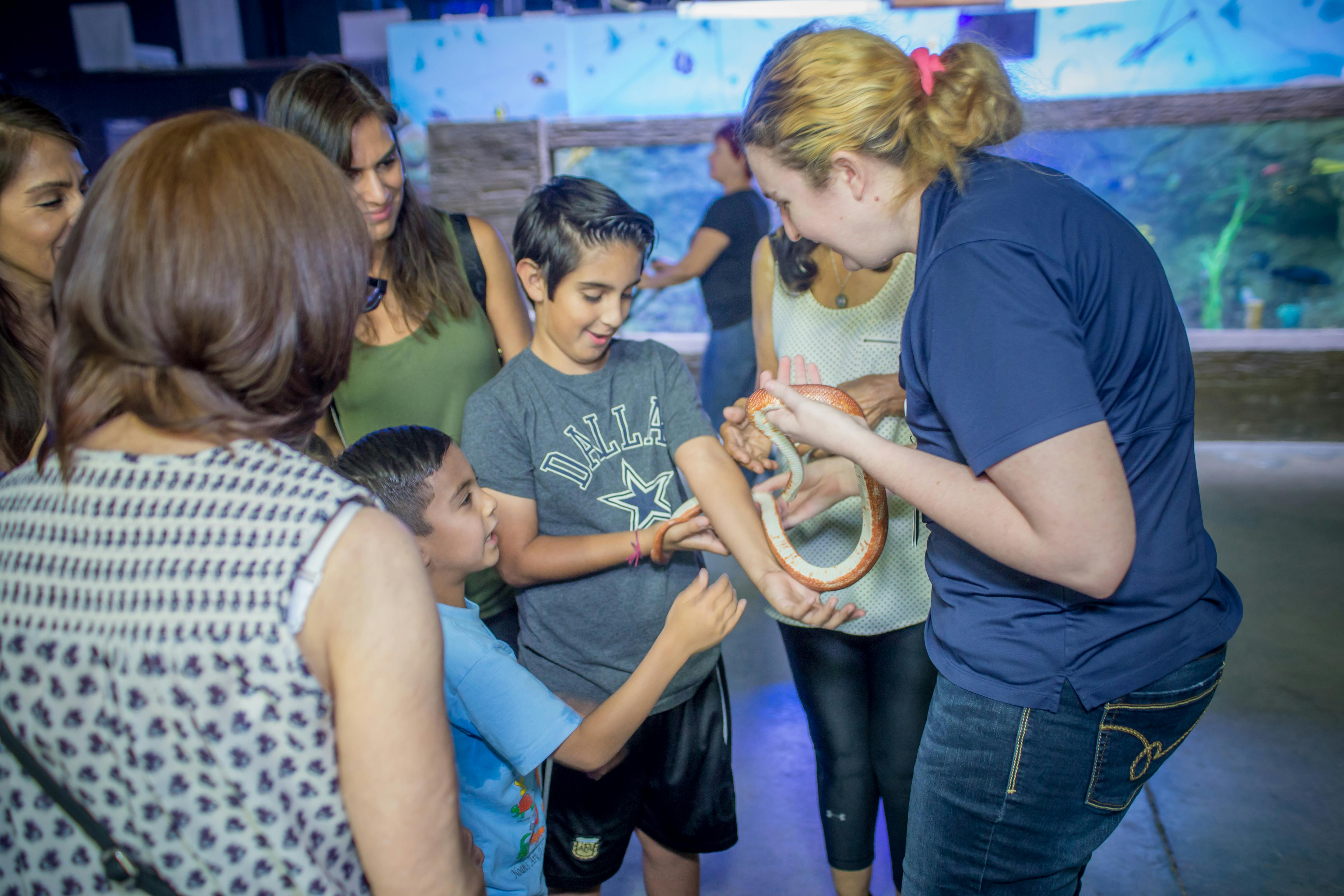 Skupina lidí, včetně dětí, interaguje se ženou, která drží hada ve vnitřním prostředí.