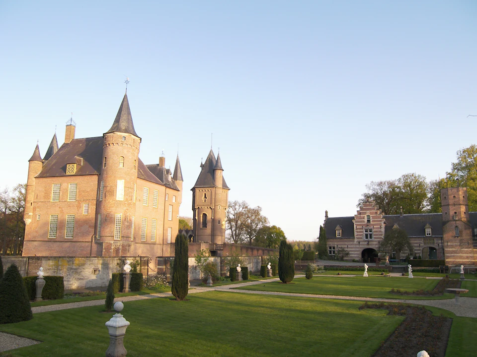 A historic castle with tall towers and a manicured garden featuring hedges and statues under a clear sky.