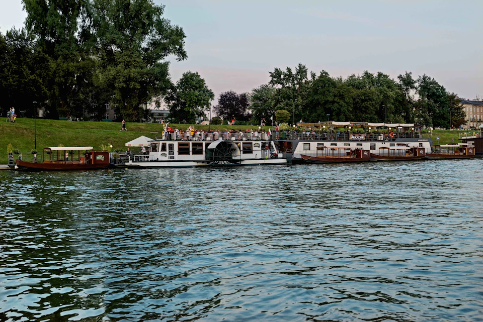 Boats are docked along a tree-lined riverbank with people walking on a grassy area. The water is calm under a light sky.