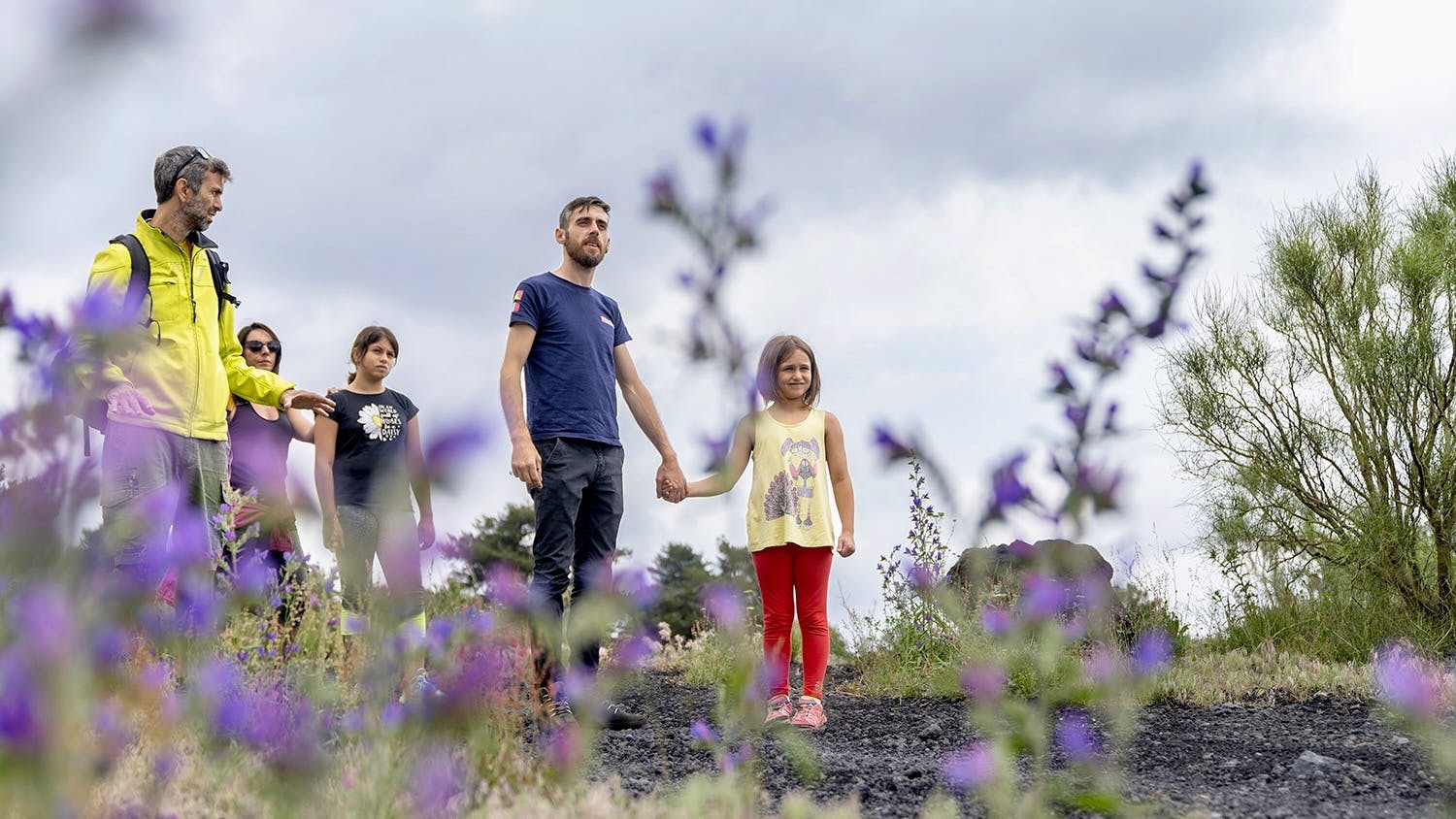 Ένας άντρας με κίτρινο μπουφάν και τρεις άλλοι, συμπεριλαμβανομένων δύο παιδιών, στέκονται σε ένα λιβάδι με μοβ λουλούδια κάτω από έναν συννεφιασμένο ουρανό.