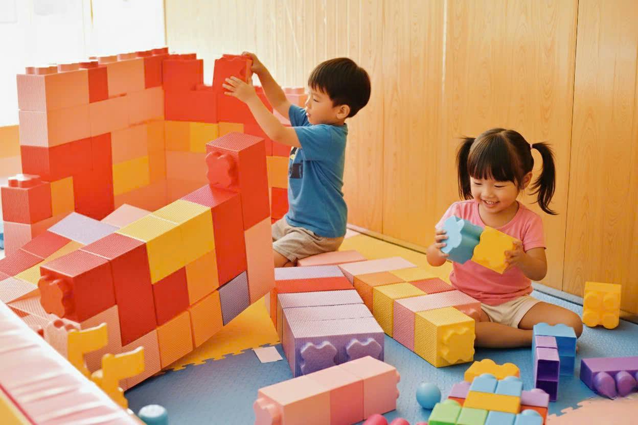 Two children playing with large colorful building blocks in a playroom with wooden walls and a soft mat floor.