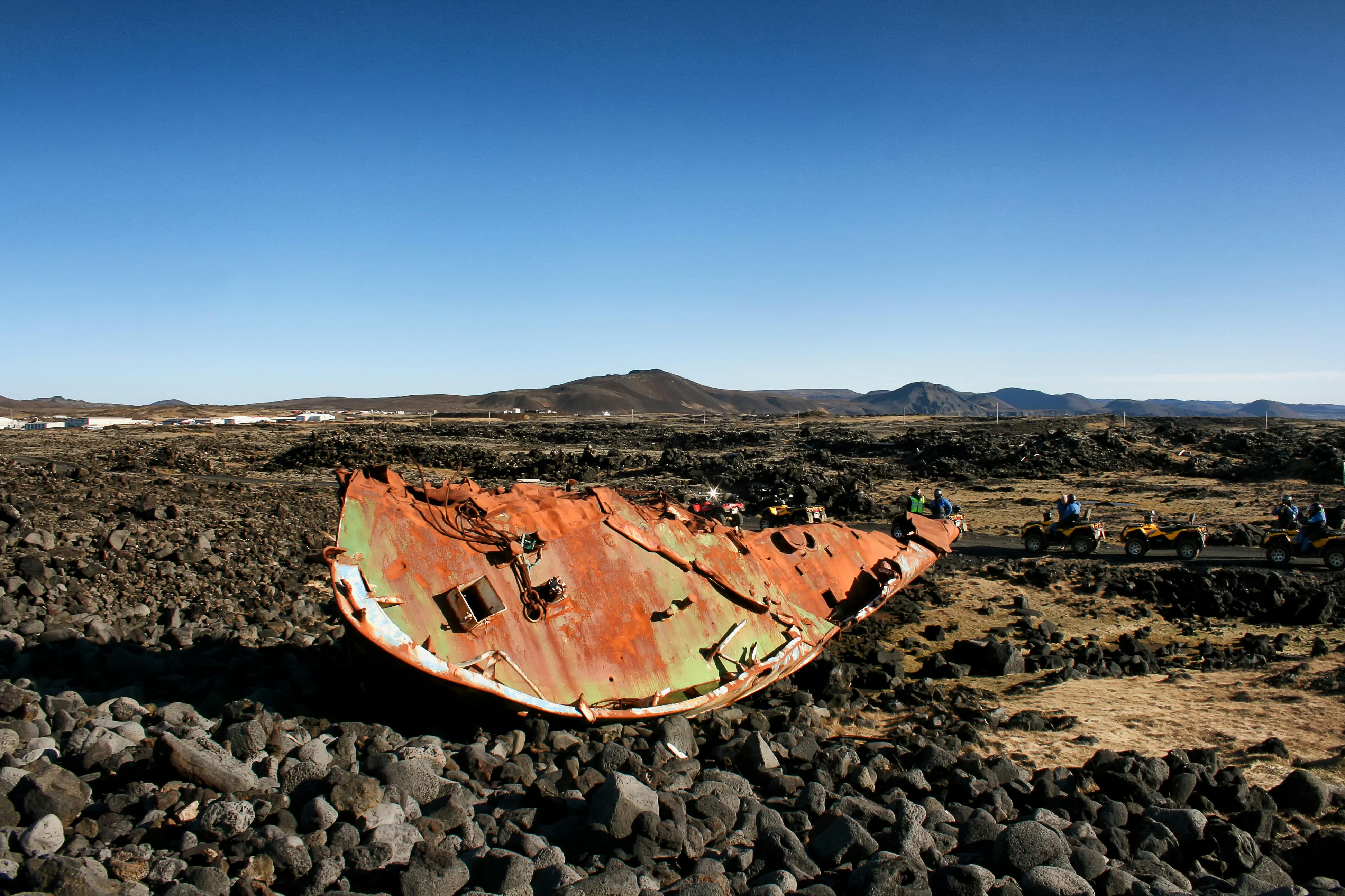 Une grande épave rouillée repose sur un paysage rocheux sous un ciel bleu clair, avec des collines et des gens au loin en arrière-plan.
