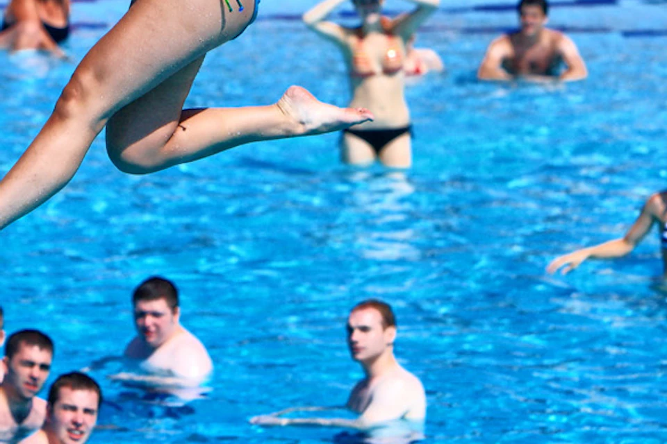 A woman in a bikini is zip-lining over a crowded pool, with people swimming and sunbathing under red umbrellas in the background.