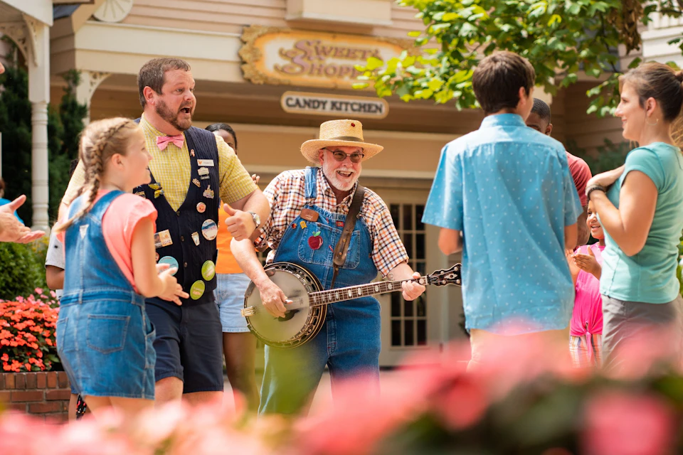A man plays a banjo outdoors near a candy shop, surrounded by smiling people, including a girl in denim overalls.