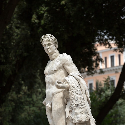 Una statua di marmo di un giovane uomo con i capelli corti e ricci drappeggiati in una tunica, davanti a degli alberi e a un edificio con finestre sullo sfondo.
