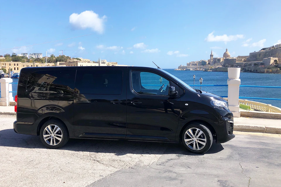 A black van is parked on a coastal road with a sea view and buildings in the background under a clear blue sky.