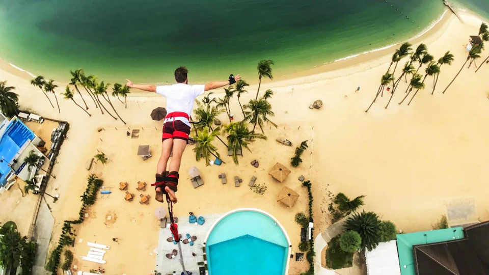 A person bungee jumping over a tropical beach with palm trees, a pool, and the ocean visible below.