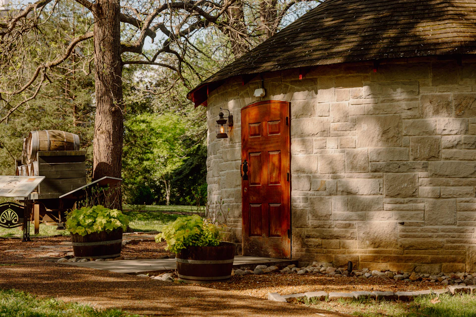 Stone building with wooden door, surrounded by trees and greenery; two wooden barrel planters with plants near the entrance.