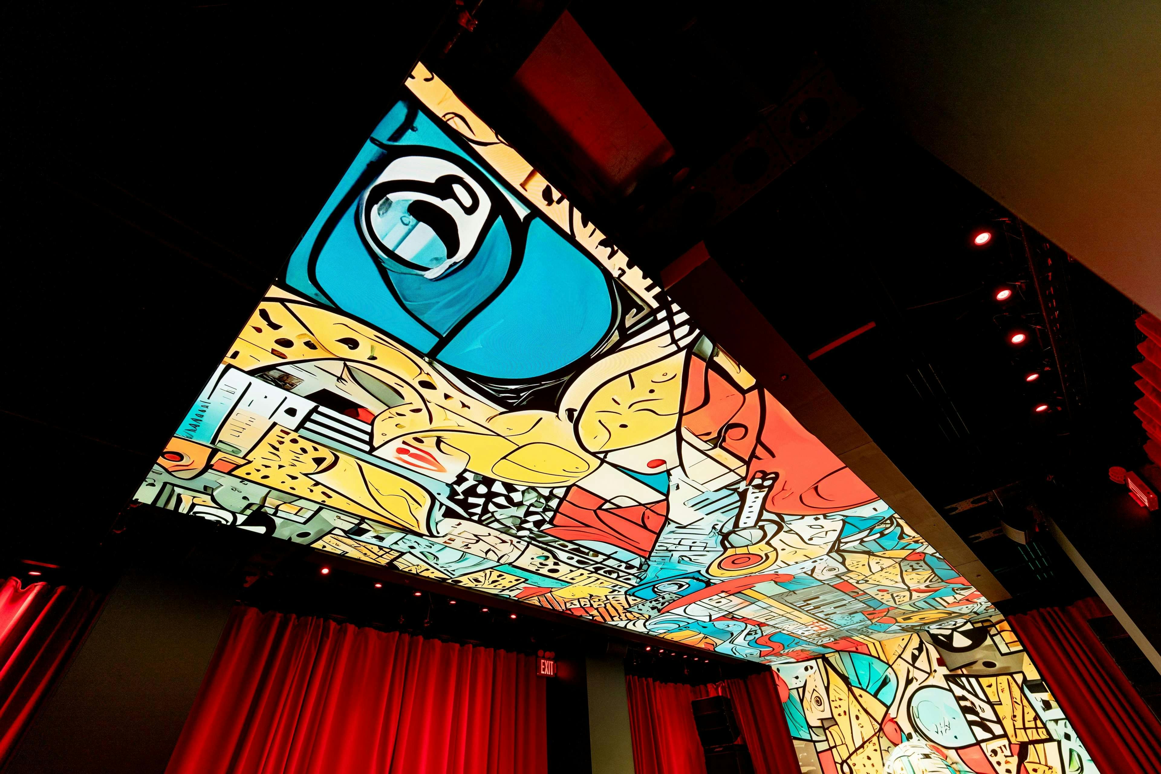 Ceiling with vibrant abstract mural featuring various shapes and colors, above a stage with red curtains and stage lights.