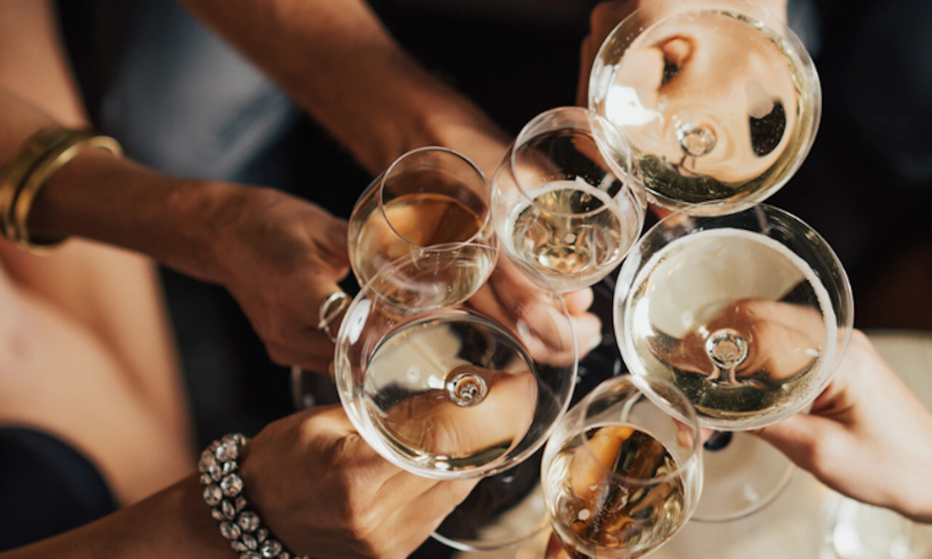 Several hands clinking various types of wine glasses filled with white wine, viewed from above against a dark background.