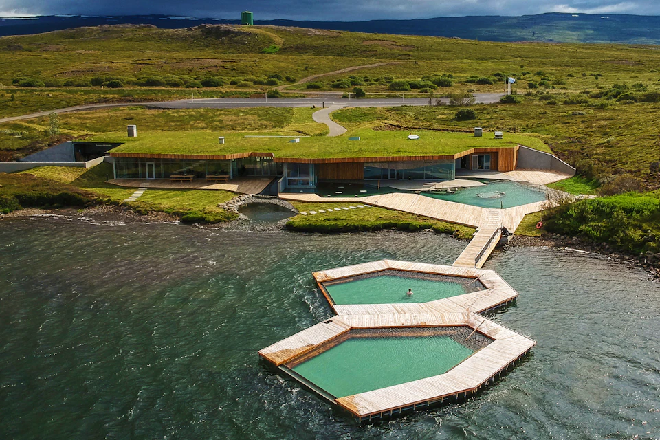 Aerial view of interconnected hexagonal pools with turquoise water, wooden walkways, and a grass-roofed building on the shore.