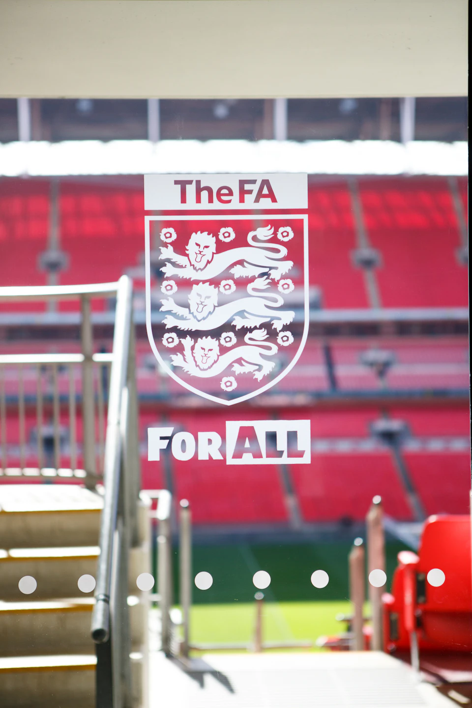 Glass panel with "The FA For All" text, three lions emblem, and red stadium seating visible through the glass.
