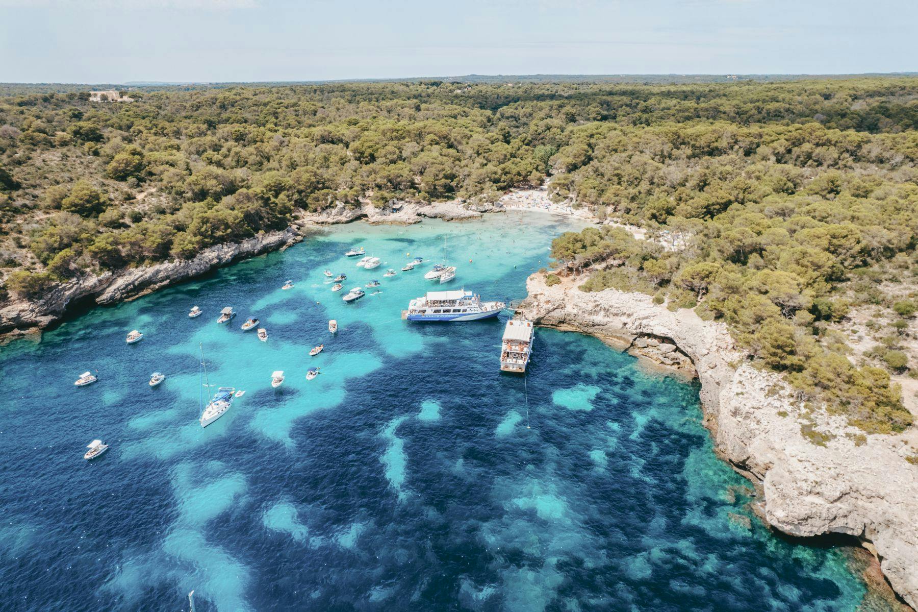 Aerial view of a bay with clear turquoise water, boats, a dock, and a forested coastline.