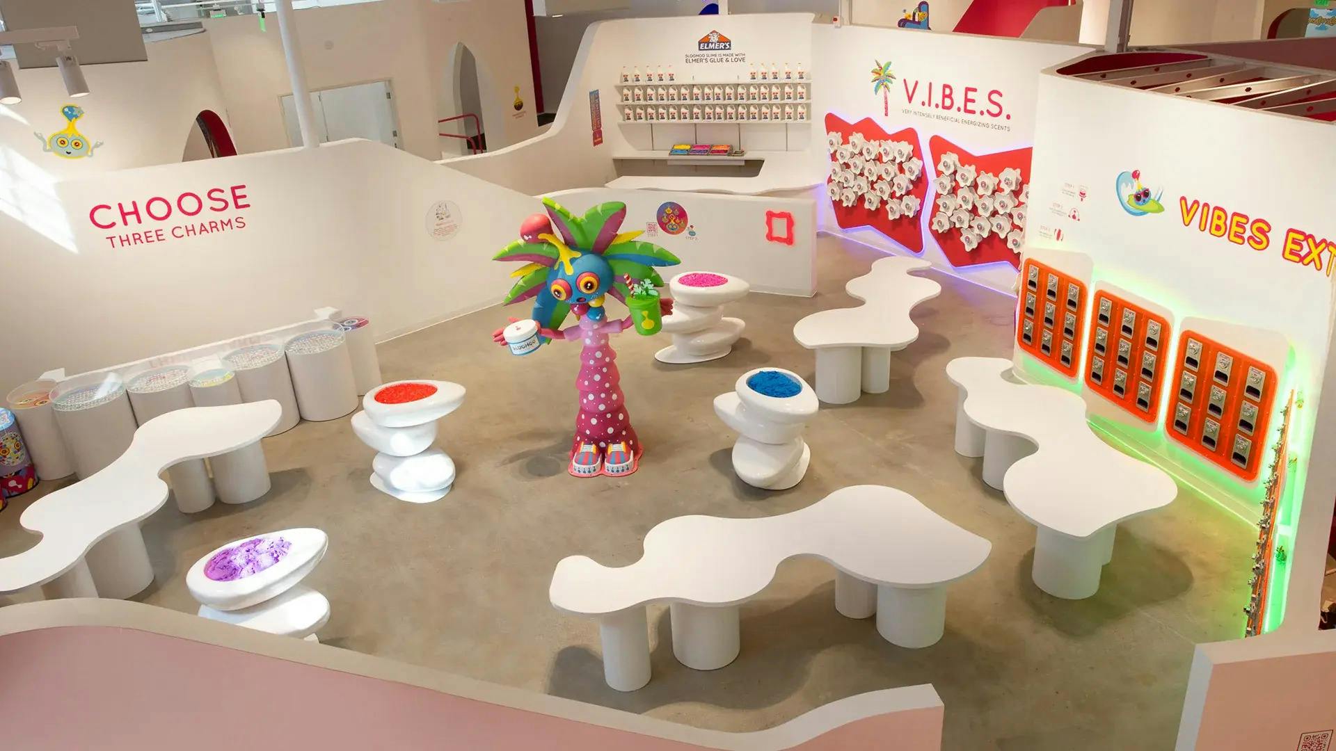 Barevná vnitřní scéna se zakřivenými bílými stoly, jedinečným posezením a hravou sochou palmy držící šálek s nápisem „V.I.B.E.S.“ na zdi.