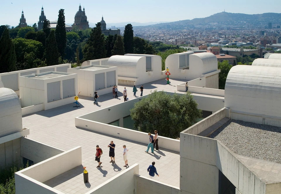 A modernist rooftop terrace featuring sculptures, people walking, and a cityscape in the background, with hills and trees visible.