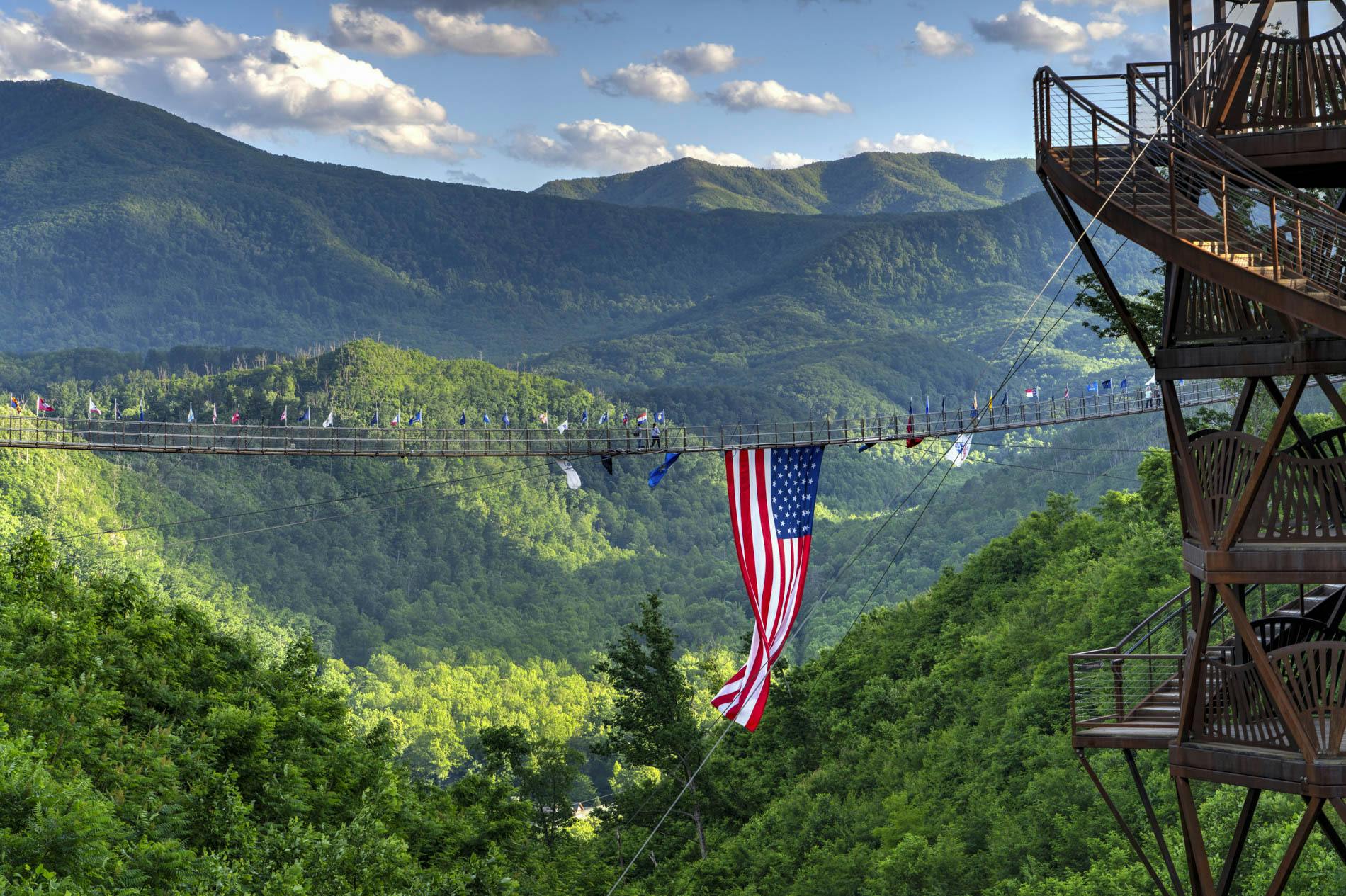 Visutý most s americkou vlajkou v zeleném zalesněném údolí s výhledem na hory v pozadí.