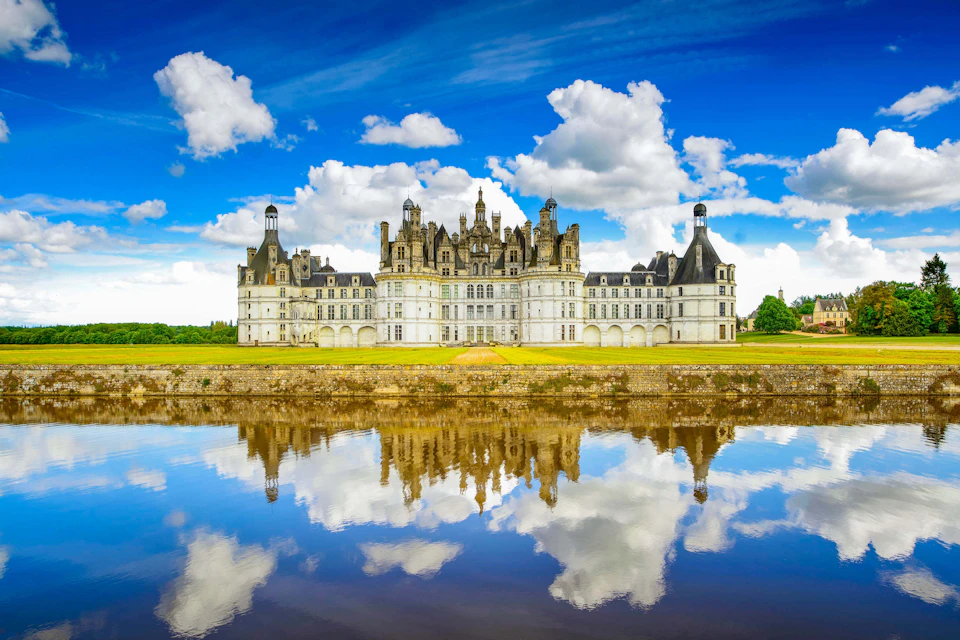 A grand castle with ornate towers and turrets, surrounded by a moat reflecting the blue sky and clouds.