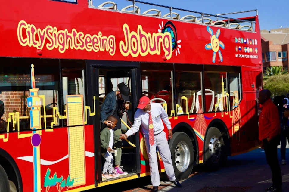 A red sightseeing bus labeled "Joburg" with a guide assisting a child and adult exiting the bus.
