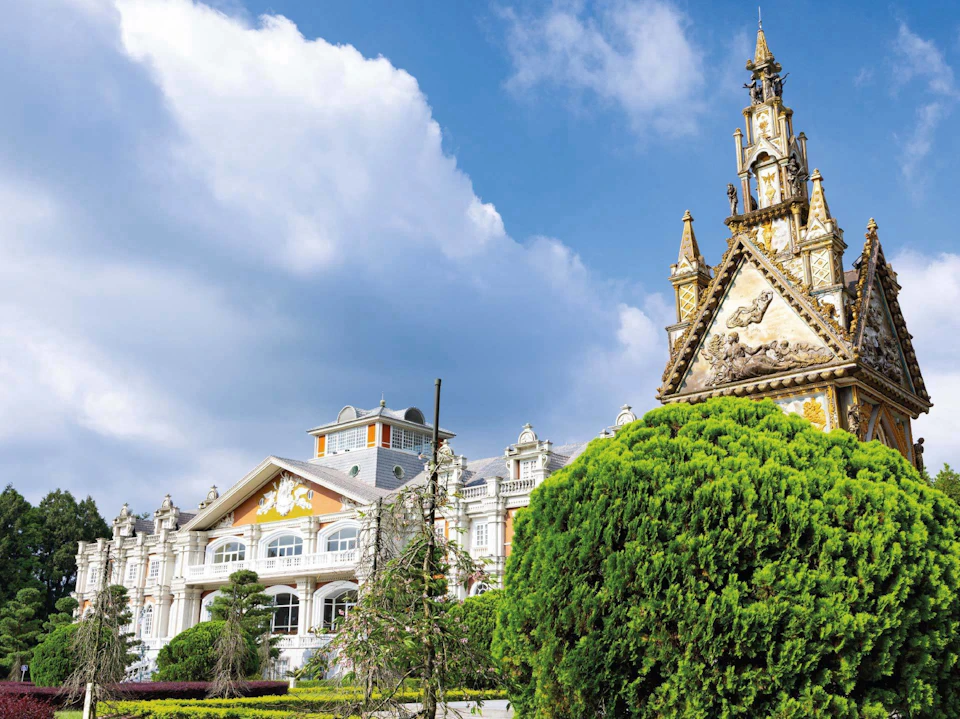 A decorative building with ornate architecture under a cloudy sky, surrounded by green trees and shrubs.
