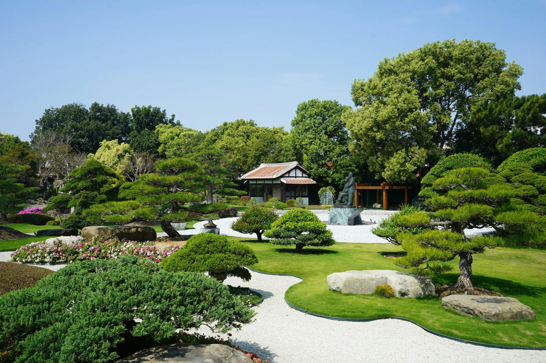 A Japanese garden with manicured trees, gravel paths, a statue, and a traditional wooden building under a clear sky.