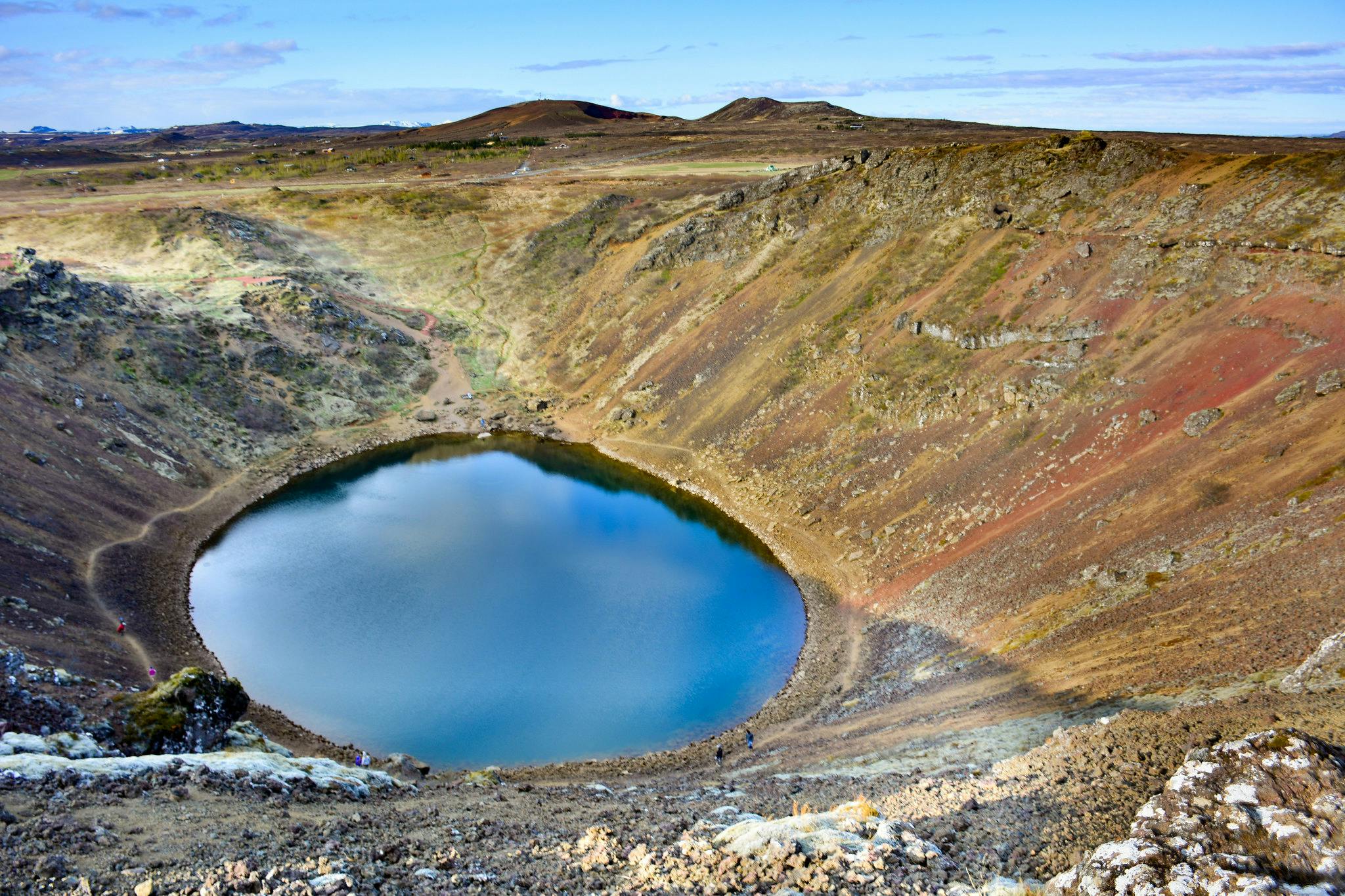 A crater lake with clear blue water surrounded by rugged, rocky terrain and sparse vegetation under a blue sky.