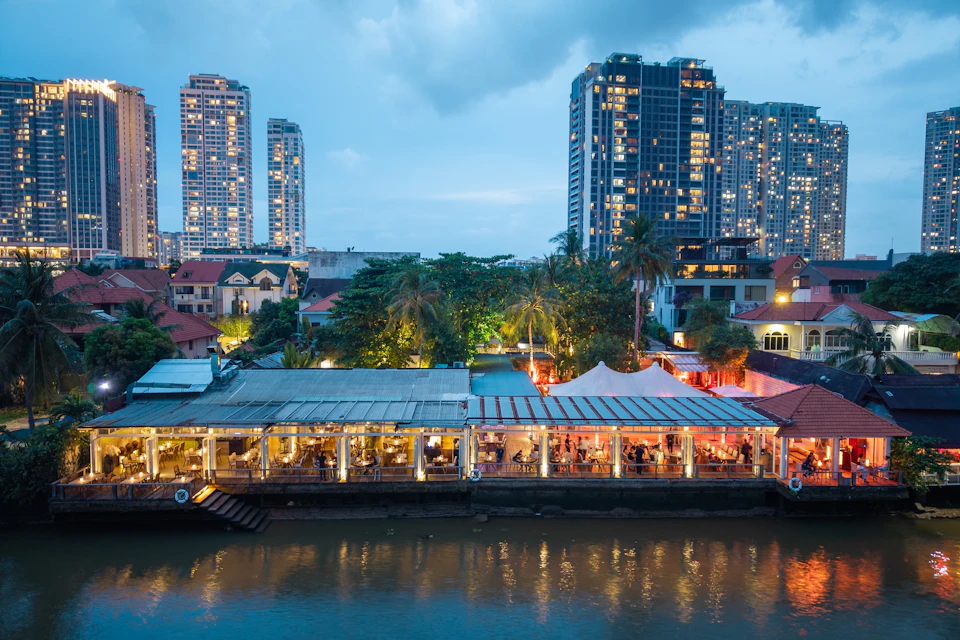 Evening view of a riverside restaurant with large windows, surrounded by trees and tall illuminated residential buildings.