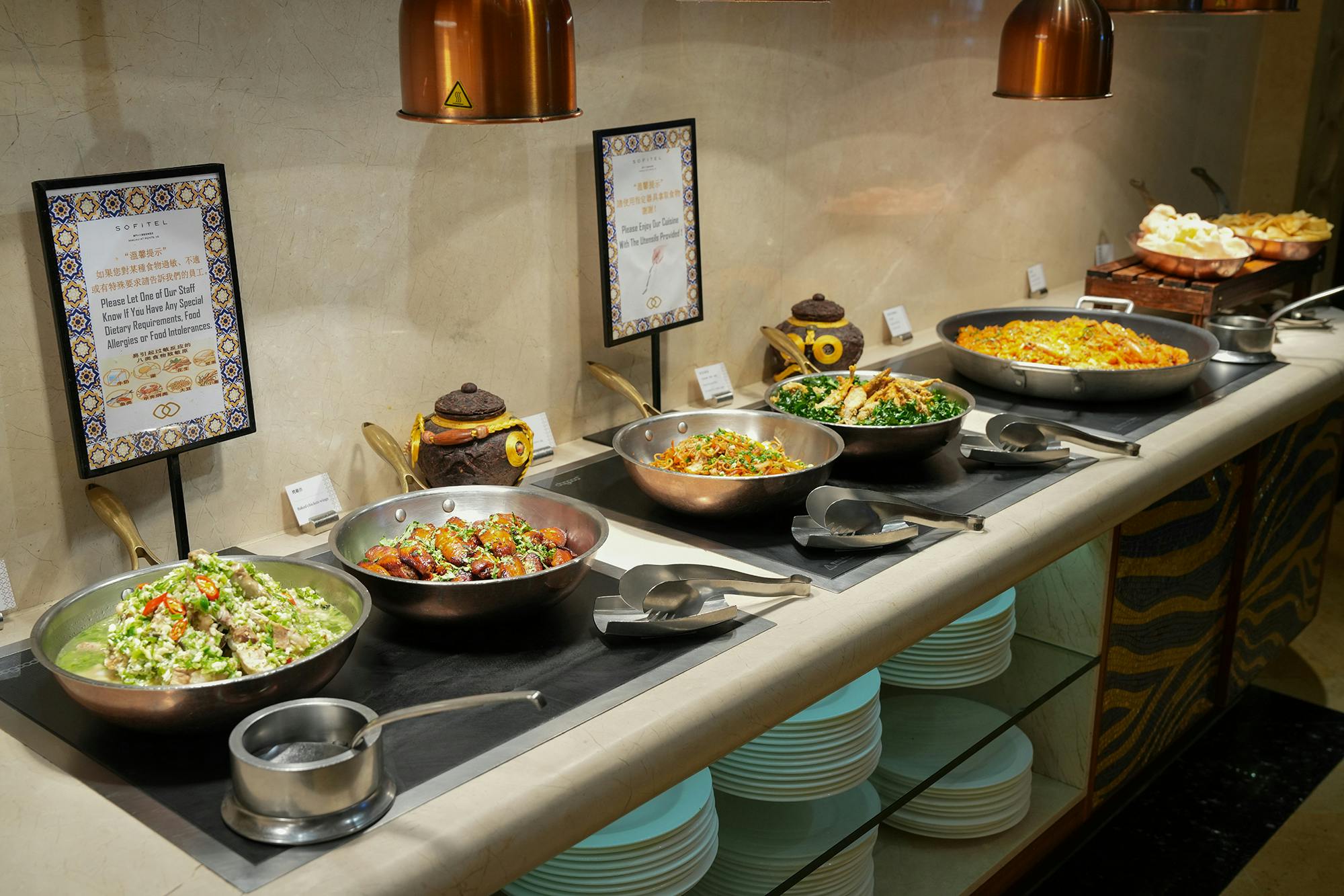 A buffet setup with various dishes in metal bowls, utensils, and plates arranged neatly under warm lighting.