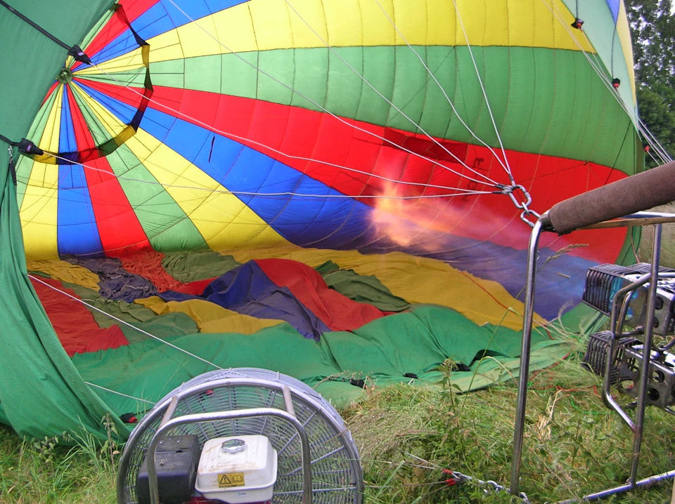 A colorful hot air balloon being inflated with a flame, connected to metal cables, with a fan in the foreground.