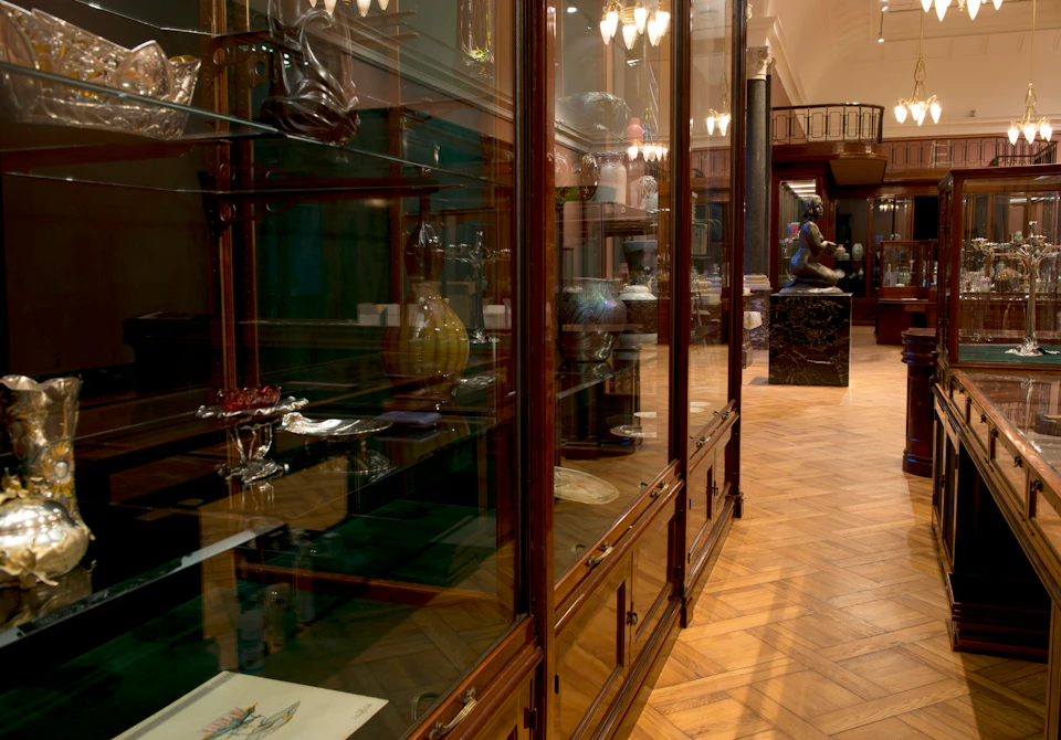 A museum hall with wooden display cases containing various artifacts and vases, and a statue visible in the background.