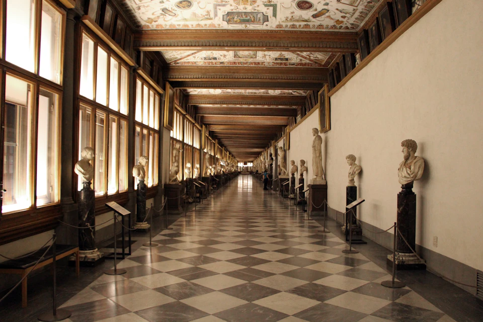 A long, ornately decorated gallery with statues and large windows lining the walls, featuring a checkered marble floor.