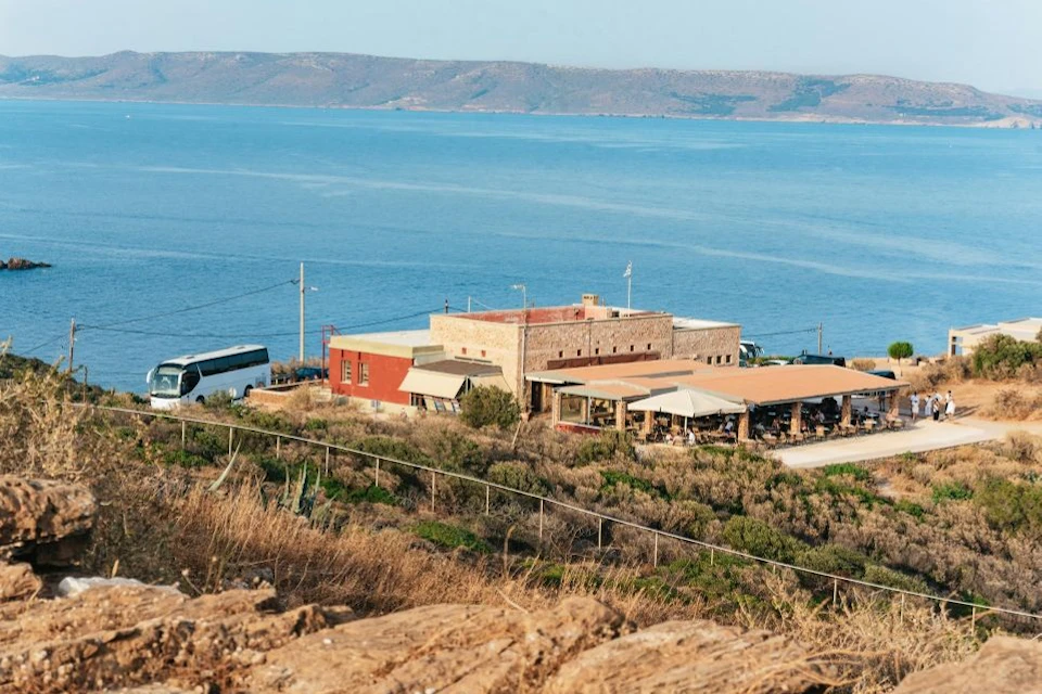 A coastal view featuring a cluster of buildings near the water, with mountains in the background and a bus parked nearby.