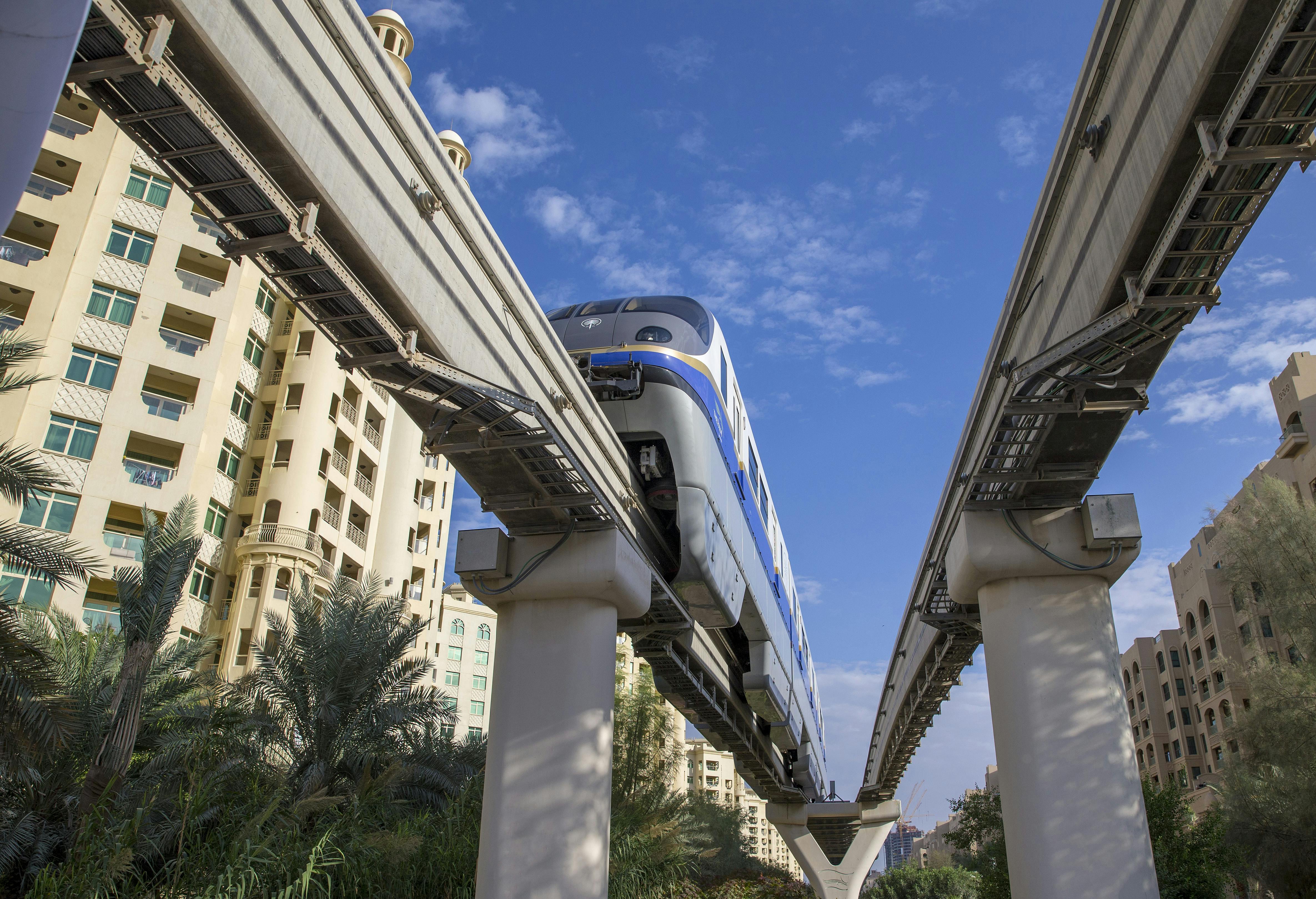 Монорельсовый поезд движется между высотными зданиями с пальмами внизу и голубым небом над головой.