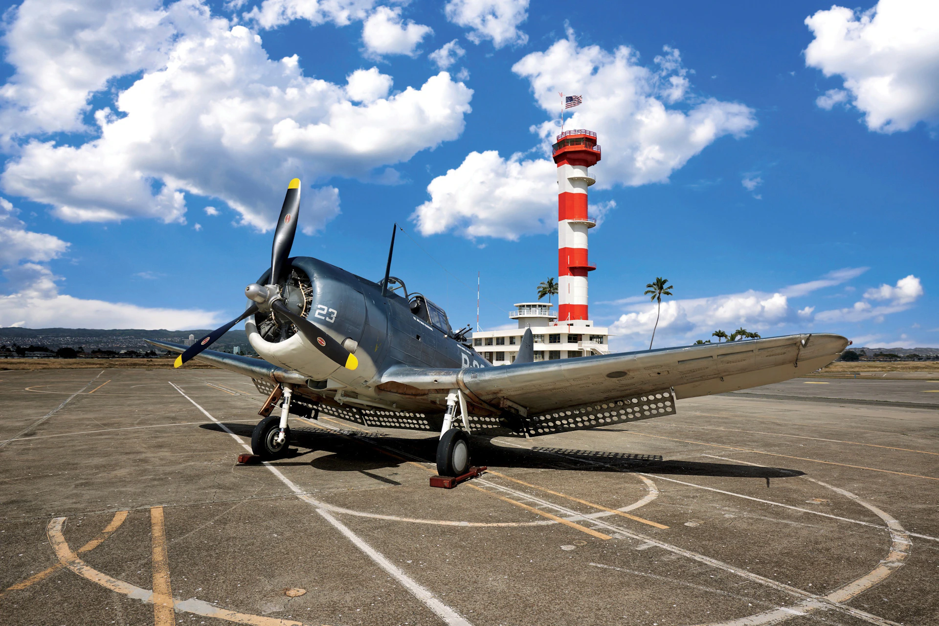 A vintage aircraft with the number 23 on its fuselage is parked on an airstrip. A red and white control tower stands in the background.