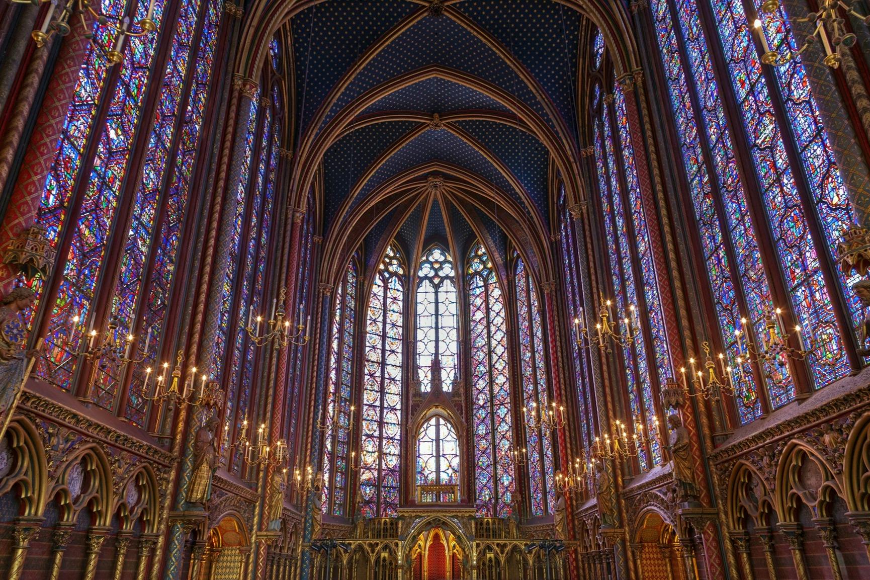 Gothic cathedral interior with vaulted ceilings, ornate stained glass windows, and chandeliers.