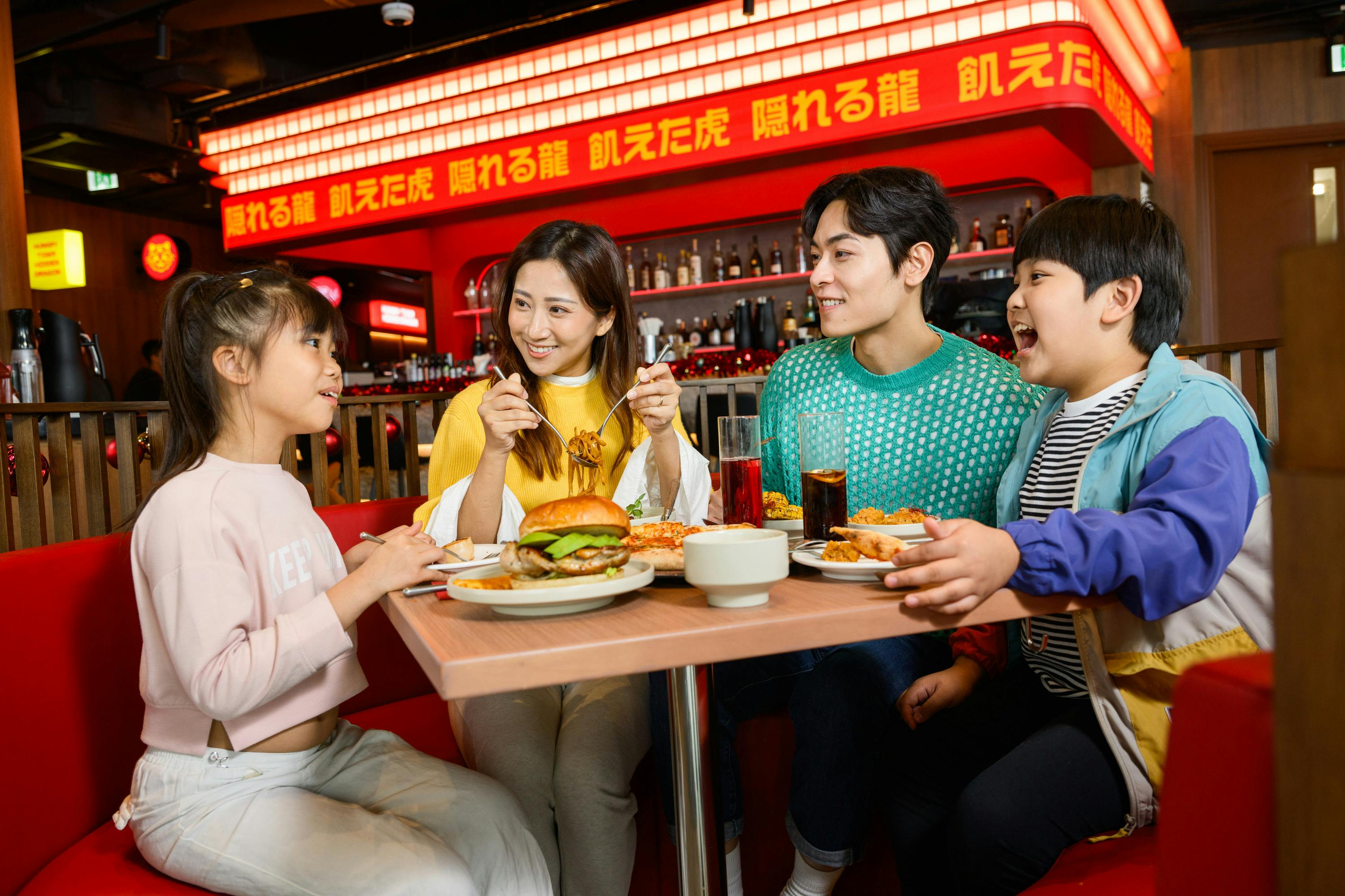 A family dining together at a restaurant with food and drinks on the table, under a red sign with text in Japanese.